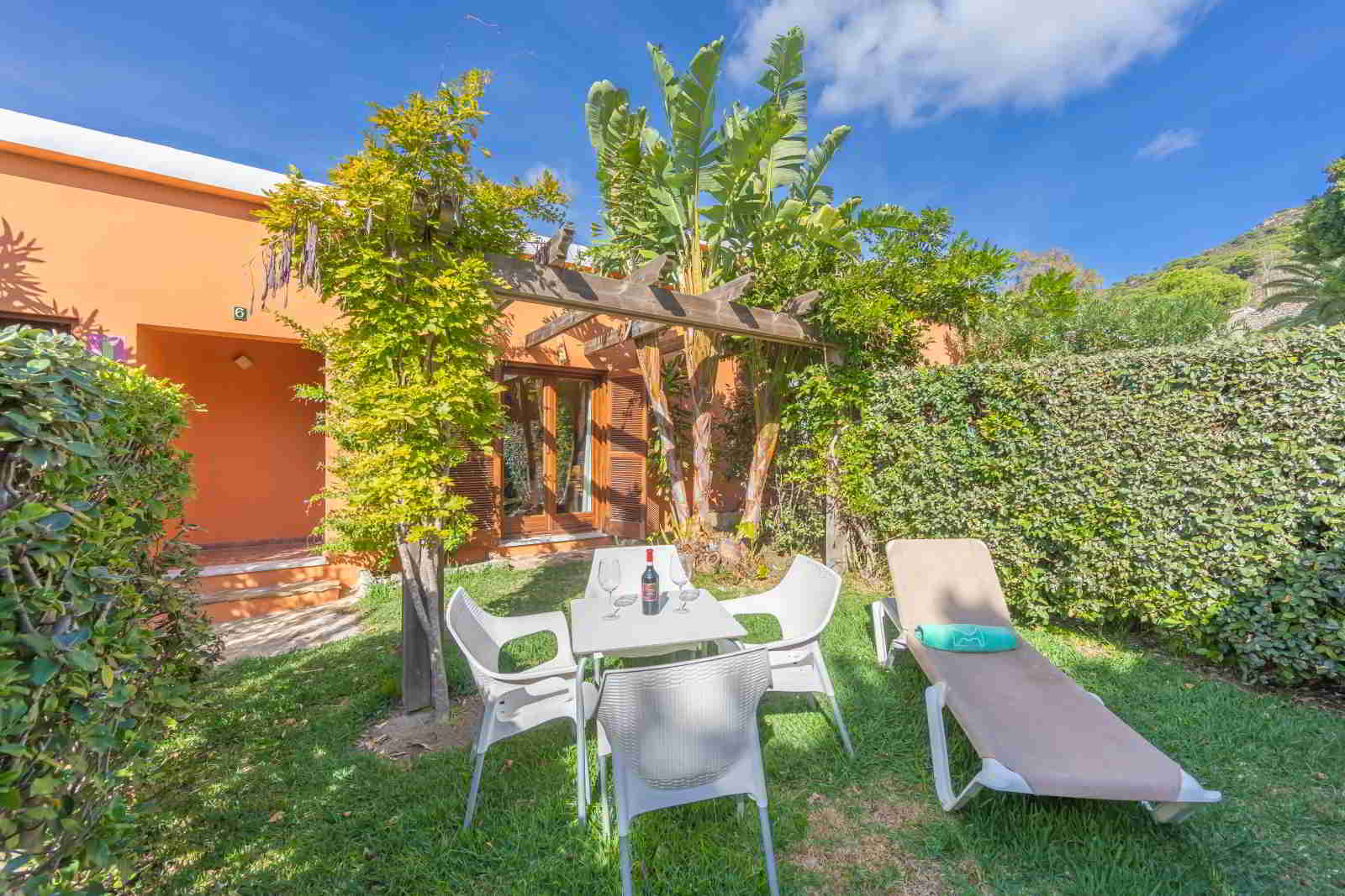 Jardín privado de habitación familiar con mesa exterior, sillas, tumbona, pérgola vegetal y entorno verde soleado.