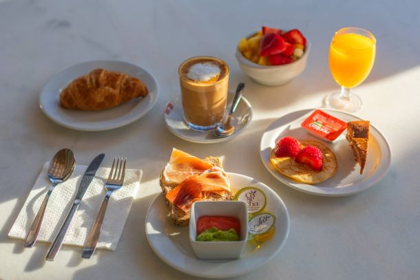Desayuno completo servido sobre mesa blanca, con zumo, café, frutas, bollería y tostadas.